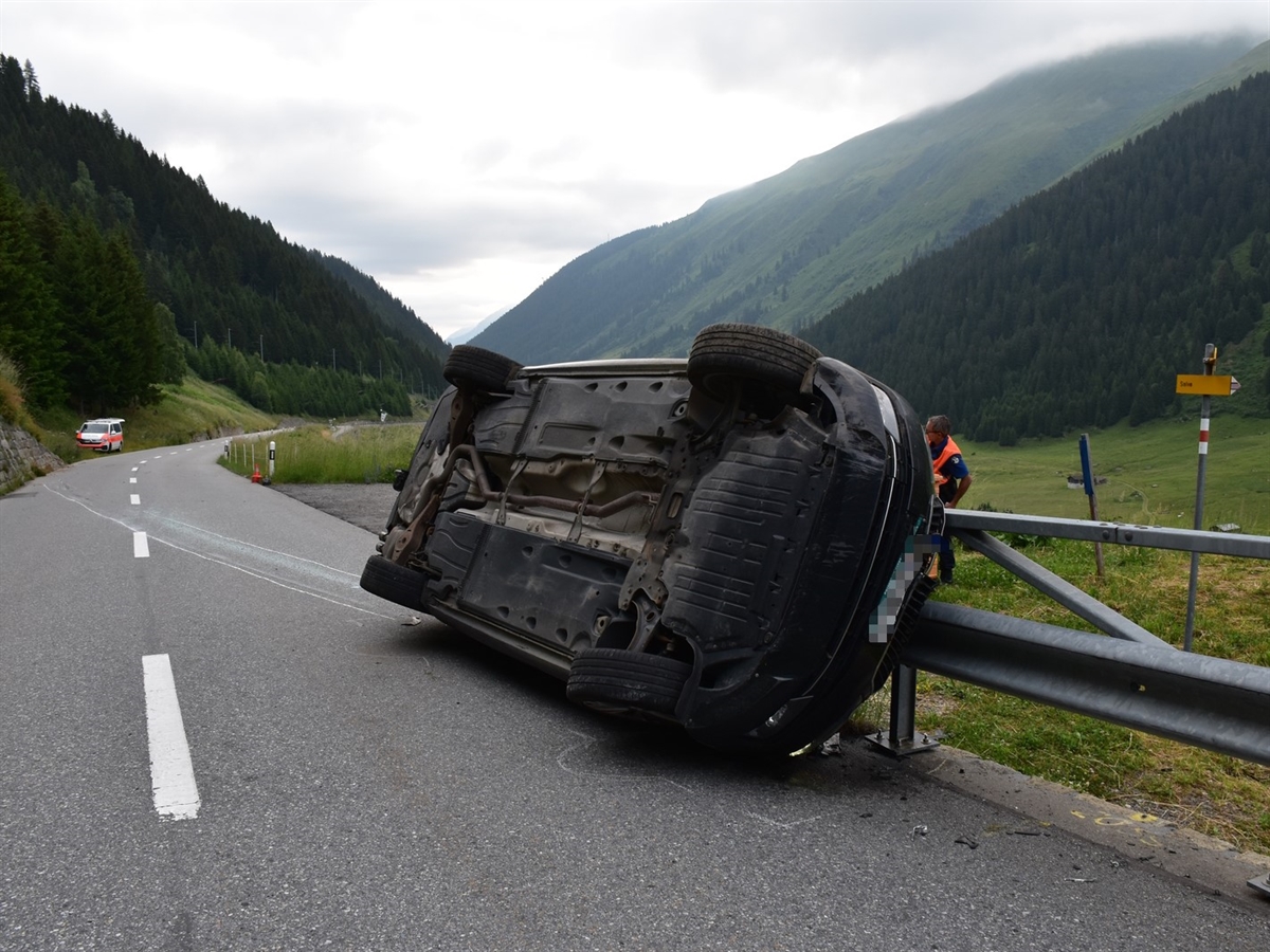 Rechts das auf der Seite liegende Auto. Links im Hintergrund ein Polizeiauto. Berge die sich vom bewölkten Horizont abgrenzen.