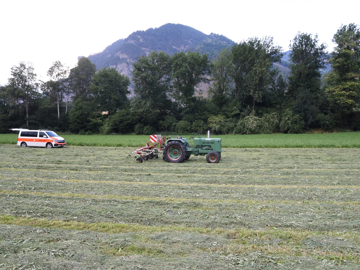 Die bewirtschaftete ebene Wiese. Im Bildzentrum ein grüner Traktor mit am Heck angebrachten Kreiselheuer. Links im Hintergrund ein Polizeiauto.