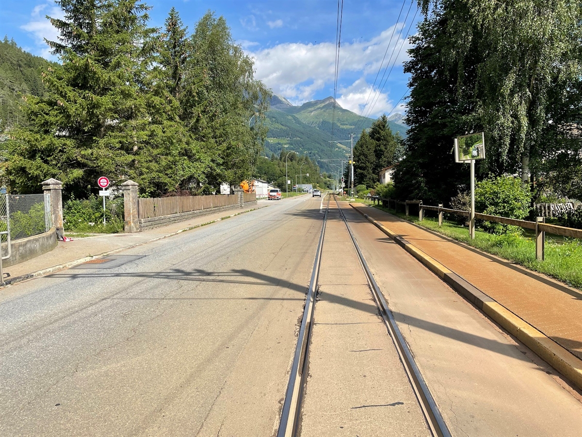 Die Strasse auf deren rechter Fahrbahnhälfte das Bahngleis eingelassen ist. Die Strasse wird beidseitig von Trottoirs von Gebäuden abgegrenzt. Im Hintergrund Berge und grösstenteils blauer Himmel.