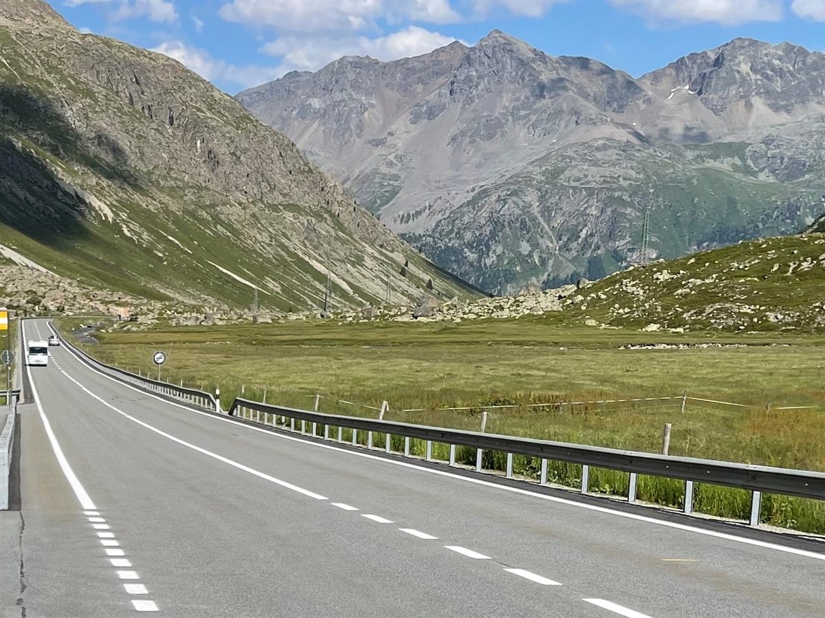 Eine Gerade, rechts davon Wiesland. Im Hintergrund Berge und leicht bewölkter Himmel.