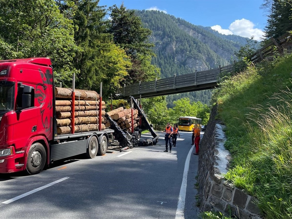 Links der Holztransporter, ein Anhängerzug. Der Anhänger ist in die Fahrbahnmitte gekippt, ebenfalls der Heckkran des Lastwagens. Rechts davon Einsatzkräfte. Im Hintergrund blauer Himmel.