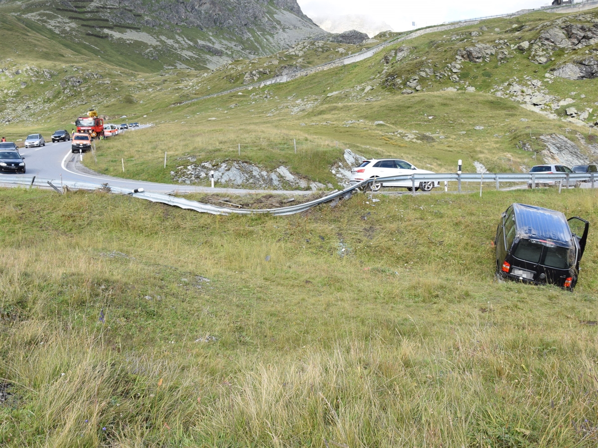 Unterhalb der Fahrbahn steht der VW-Bus auf dem Wiesland. Die Leitplanke ist völlig aus der Verankerung gerissen und liegt ebenfalls neben der Fahrbahn. Mehrere Autos fahren an der Unfallstelle vorbei. Ein Fahrzeug des Tiefbauamtes Graubünden sowie ein Abschleppfahrzeug stehen oberhalb der Unfallstelle.
