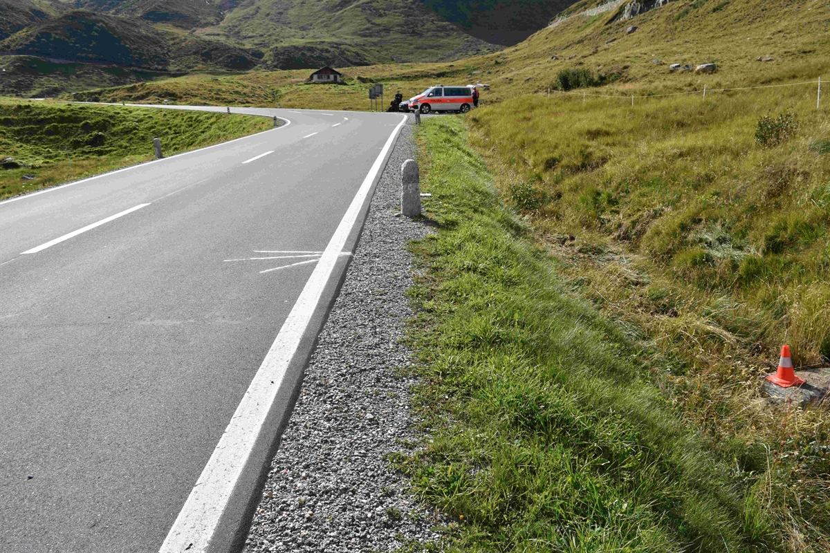 Es ist Tag und die Strasse ist trocken. Rechts neben der Strasse im Wiesland steht ein Molankegel als Unfallmarkierung und im Hintergrund auf einem Abstellplatz das Polizeiauto mit dem Unfallmotorrad.