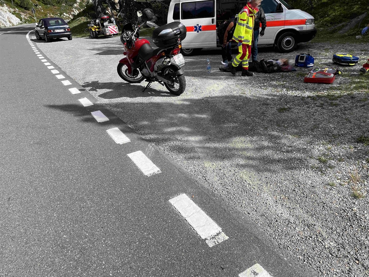 Es ist Tag und die Strasse ist trocken. Auf dem rechtsseitigen unbefestigten Abstellplatz stehen das Unfallmotorrad und seitlich im Hintergrund die Ambulanz sowie die Rettungsperson und die Sozia liegt am Boden.
