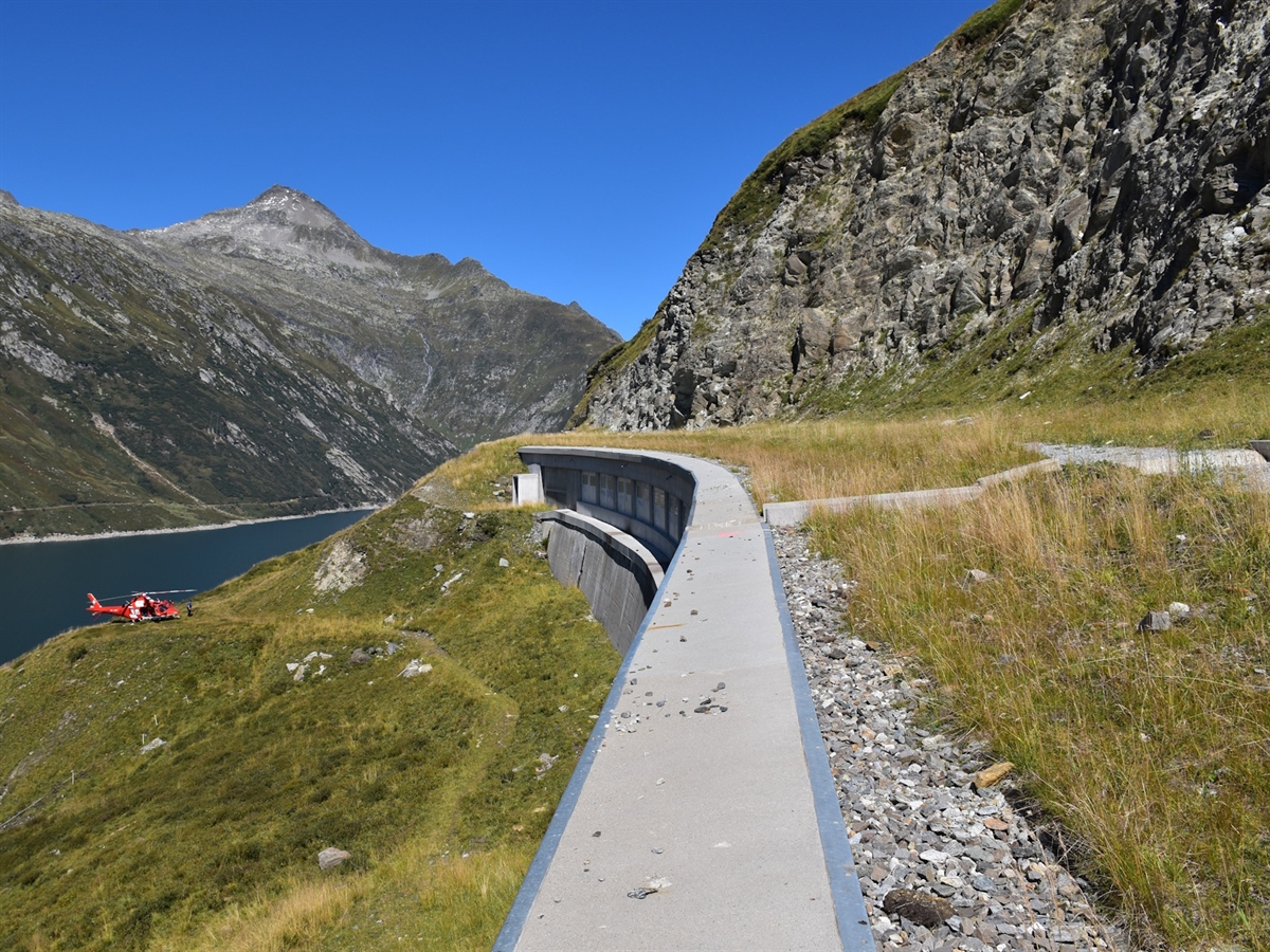 In Bildmitte die Betonbrüstung, rechts das Galeriedach und links tiefer gelegenes Wiesland. Ein Rega-Helikopter, der Stausee. Im Hintergrund strahlend blauer Himmel und der Blick auf Bündner Berge.