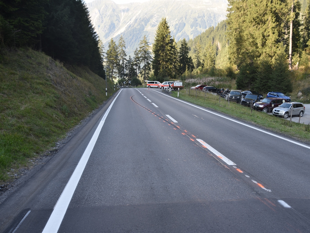 Eine Blockier-/Bremsspur des Motorrads die auf die Gegenfahrspur führt. Einsatzfahrzeuge der Polizei und weitere Fahrzeuge auf einem Parkplatz rechts der Strasse. Im Hintergrund Bäume.
