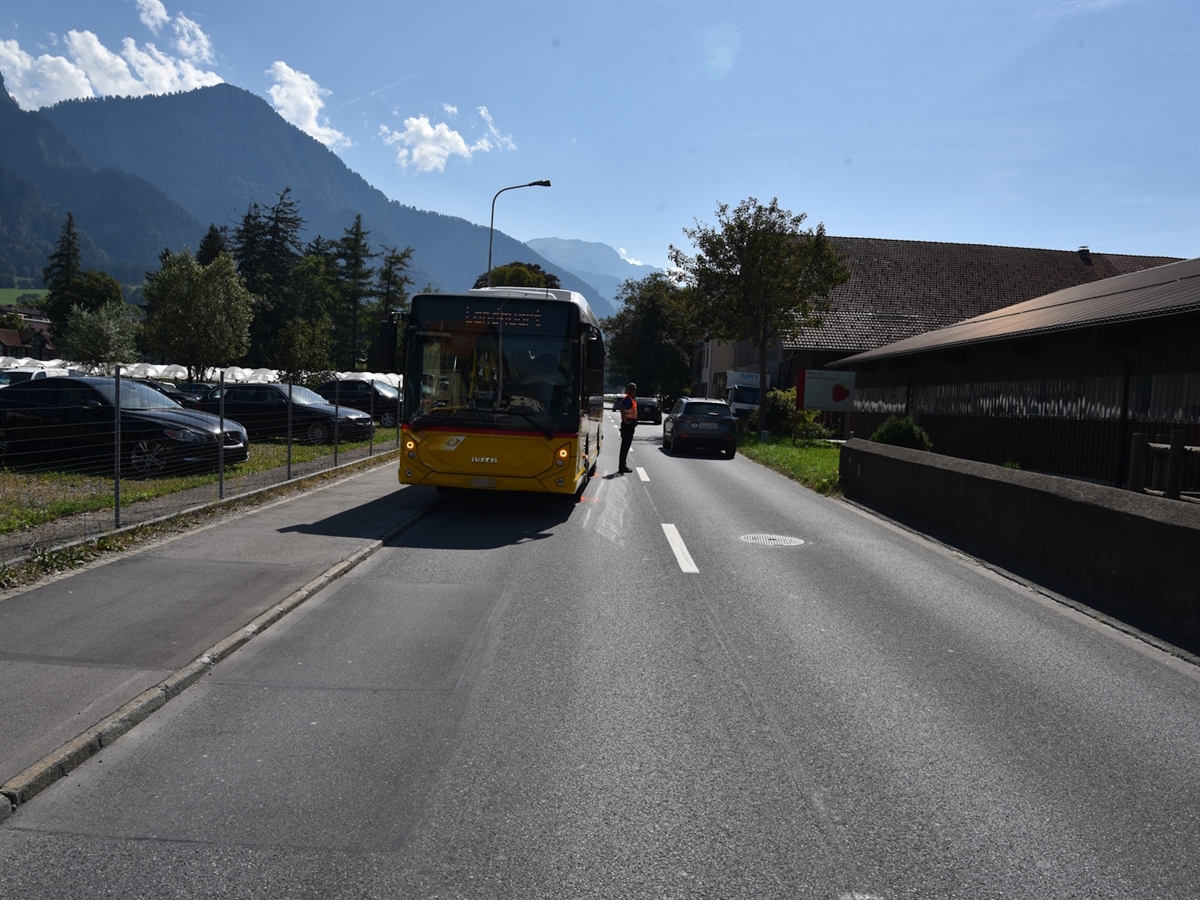 Links das Trottoir, rechts davon das Postauto und ein Polizist. Rechts ein Gebäude, im Hintergrund Berge und der Horizont.