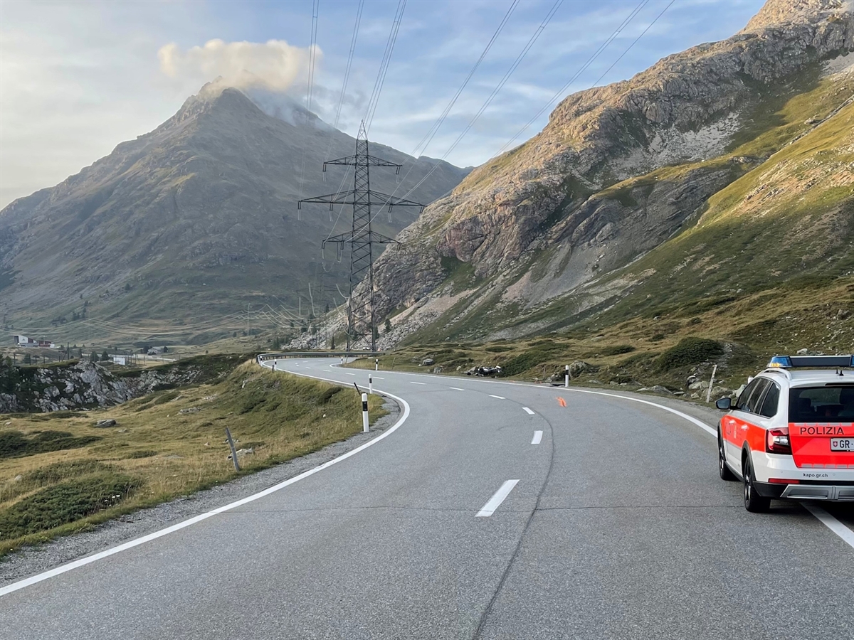Rechts ein Polizeiauto. Links davon die Unfallkurve mit einer eingezeichneten Spur. In der Flucht der Spur liegt das Motorrad in der Böschung. Im Hintergrund ein Berg und Horizont.