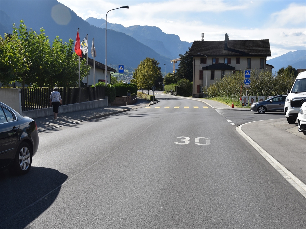Blick auf der Hauptstrasse in Richtung Igis. Von rechts mündet die Zollstrasse ein. Im Hintergrund Berge und leicht bewölkter Horizont.