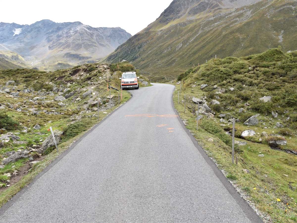 Die von alpinem Wiesland umsäumte Strasse. Rechts auf dieser Unfallspuren. Links ein Polizeiauto. 
