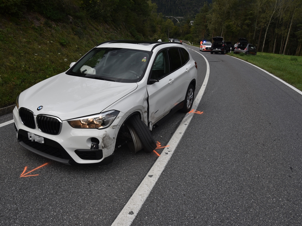 Frontal ein weisses Auto mit vorne links total beschädigter Radaufhängung. Im Hintergrund das zweite Unfallauto und ein Polizeiauto. 