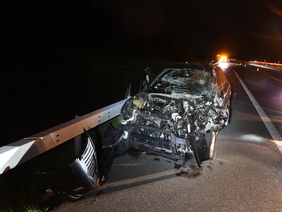 Das sehr stark beschädigte schwarze Auto steht auf dem Pannenstreifen. Es liegen Teile auf dem Boden. Die komplette Haube ist entfernt, die Scheibe zerrissen und alles ist eingedrückt.