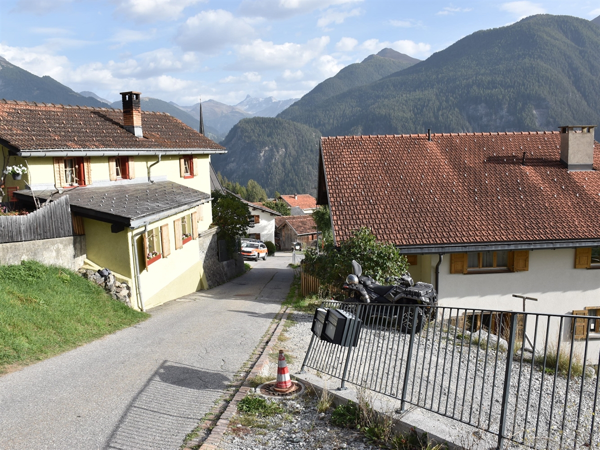 Die ins Dorf führende schmale Strasse. Zwei Gebäude begrenzen die Fahrbahn. Auf der rechten Seite ein beschädigter Eisenzaun. Hinter diesem steht das Unfallfahrzeug.
