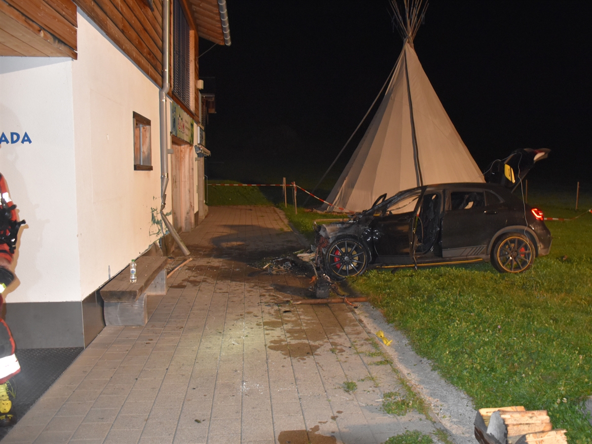 Links die Hausmauer mit einem beschädigten Dachkennel. Rechts davor steht das verunfallte Auto auf der Wiese. Hinter diesem steht ein Tipi-Zelt. Links ragt noch ein Arm eines Feuerwehrmannes ins Bild.