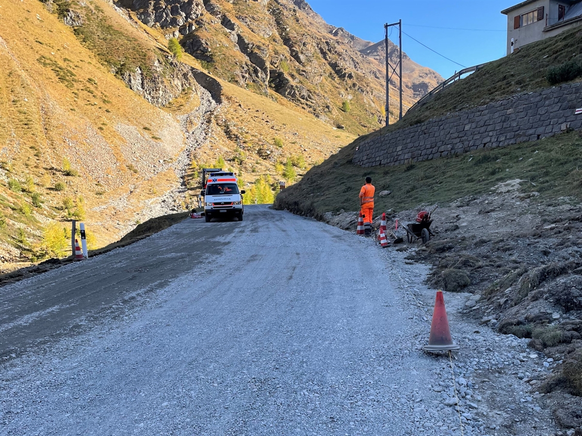 Schotterfahrbahn mit herbstlichen Bergen im Hintergrund. Auf der Fahrbahn stehen ein Rettungswagen sowie ein Arbeiter. Das Bild wird mit einem strahlend blauen Himmel abgerundet.