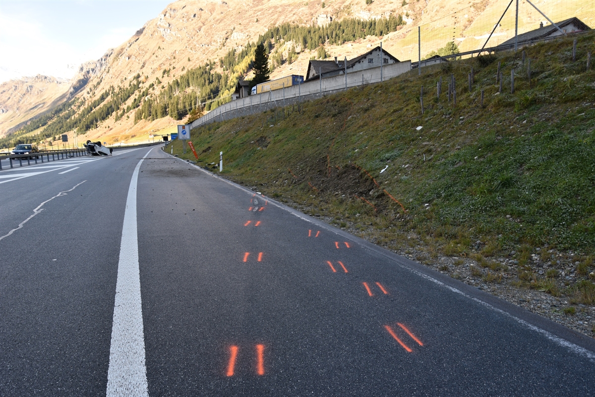 Fahrbahn mit rechtsseitig ansteigender Böschung inkl. Spuren des Unfallfahrzeuges. Total beschädigter Pw liegt im Hintergrund auf dem Dach auf der Fahrbahn.