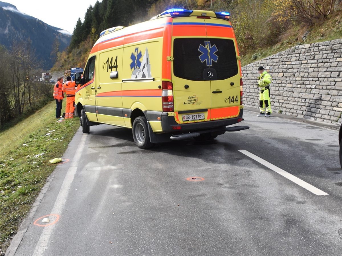Die Unfallstelle mit einem Rettungswagen der Rettung Surselva mitten im Bild. Ein Polizist spricht mit einem Bauarbeiter. Neben einer Stützmauer steht ein Verkehrsdienstspezialist.