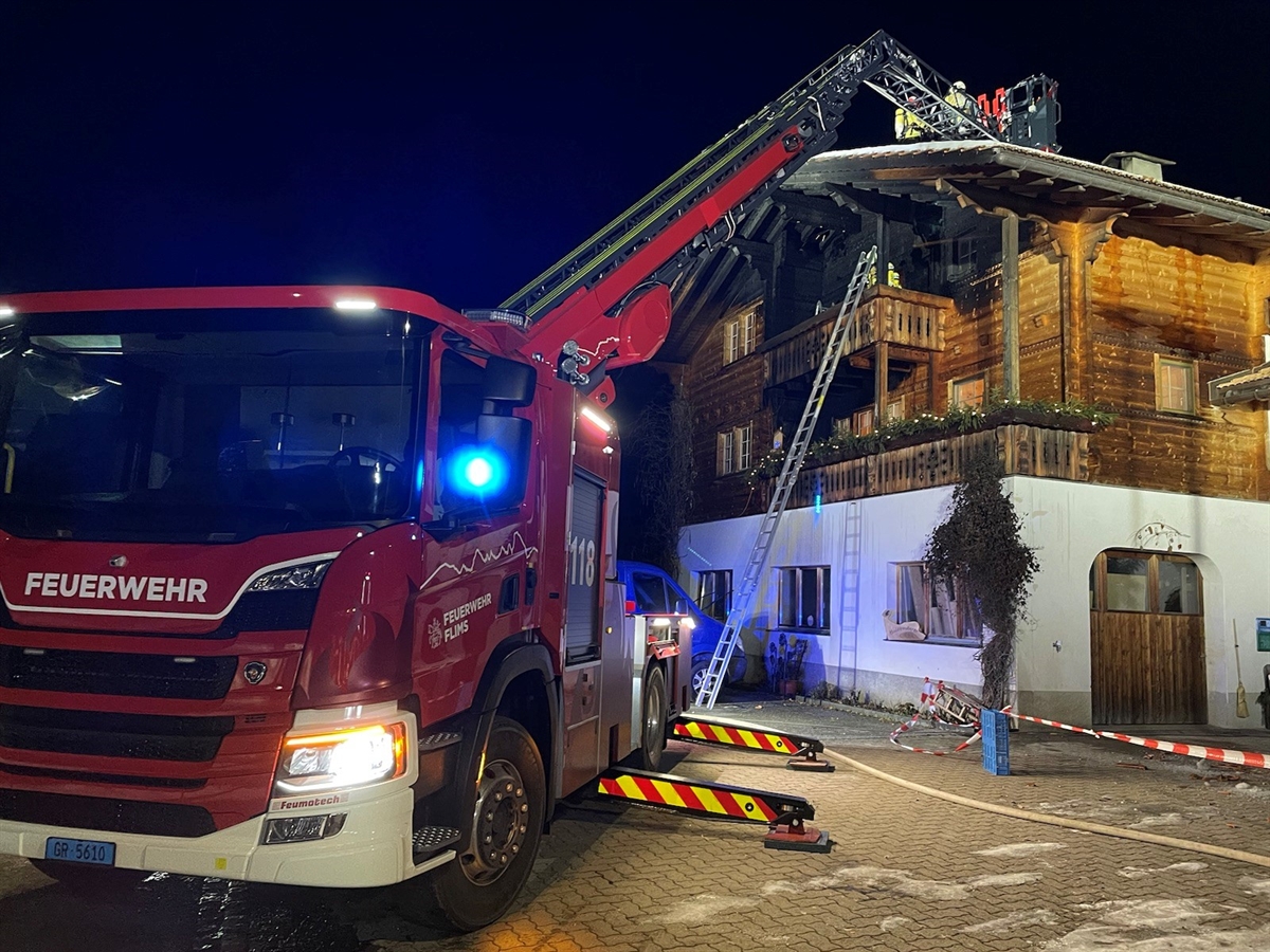 Im Vordergrund links ein Feuerwehrauto. Dessen Drehleiter ist nach rechts auf das Dach des an der Holzfassade beschädigten gerichtet. Einsatzkräfte auf dem Hausdach.