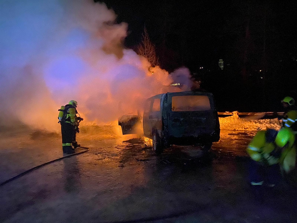 Das im Frontbereich brennende Fahrzeug von schräg hinten. Links und rechts davon Einsatzkräfte. 