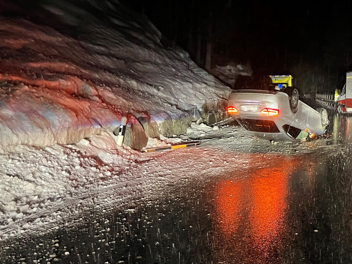 Die steil ansteigende Böschung mit Spuren des Autos. Dieses liegt auf der Fahrbahn auf dem Dach. Hinter dem Auto steht ein Rettungswagen. Rechts am Fahrbahnrand ist die Front eines parkierten Lieferwagens ersichtlich.
