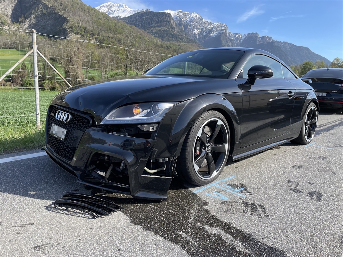 Schadenaufnahme des Autos von schräg vorne links. Kühlflüssigkeit am Boden. Im Hintergrund Berge und blauer Himmel.p