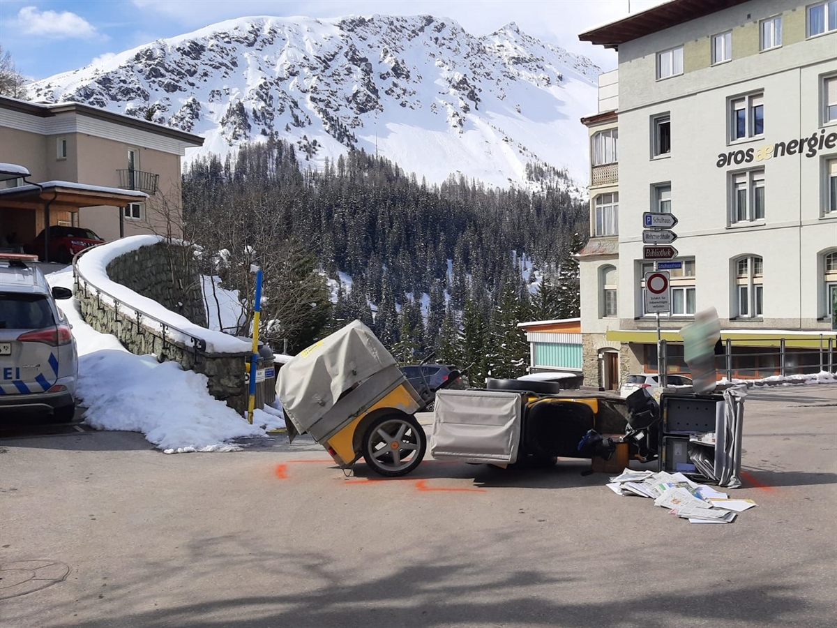Winterstimmung. Im Hintergrund ein Berg, links ein Polizeiauto und rechts davon das umgeworfene dreirädrige Zustellfahrzeug. Auf dem Boden liegen Zeitungen und Briefe.