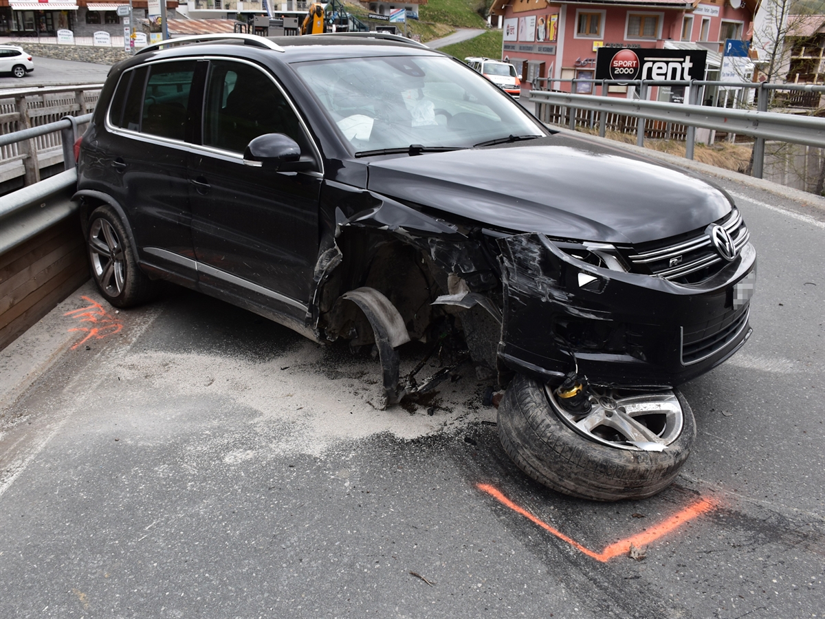Beim Unfallauto wurde das ganze vordere Rad rechts abgerissen. Das Auto steht quer zur Fahrbahn auf der Brücke. Im Hintergrund ein Polizeifahrzeug sowie Häuser von Samnaun Dorf