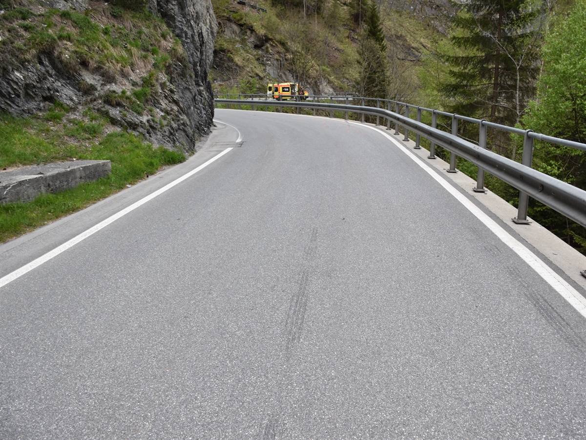 Die Linkskurve der Valserstrasse. In der Verlängerung der Kurve steht ein Ambulanzfahrzeug. Die Fahrbahn wird links und rechts vom saftige Grün der Bündner Wälder flankiert.
