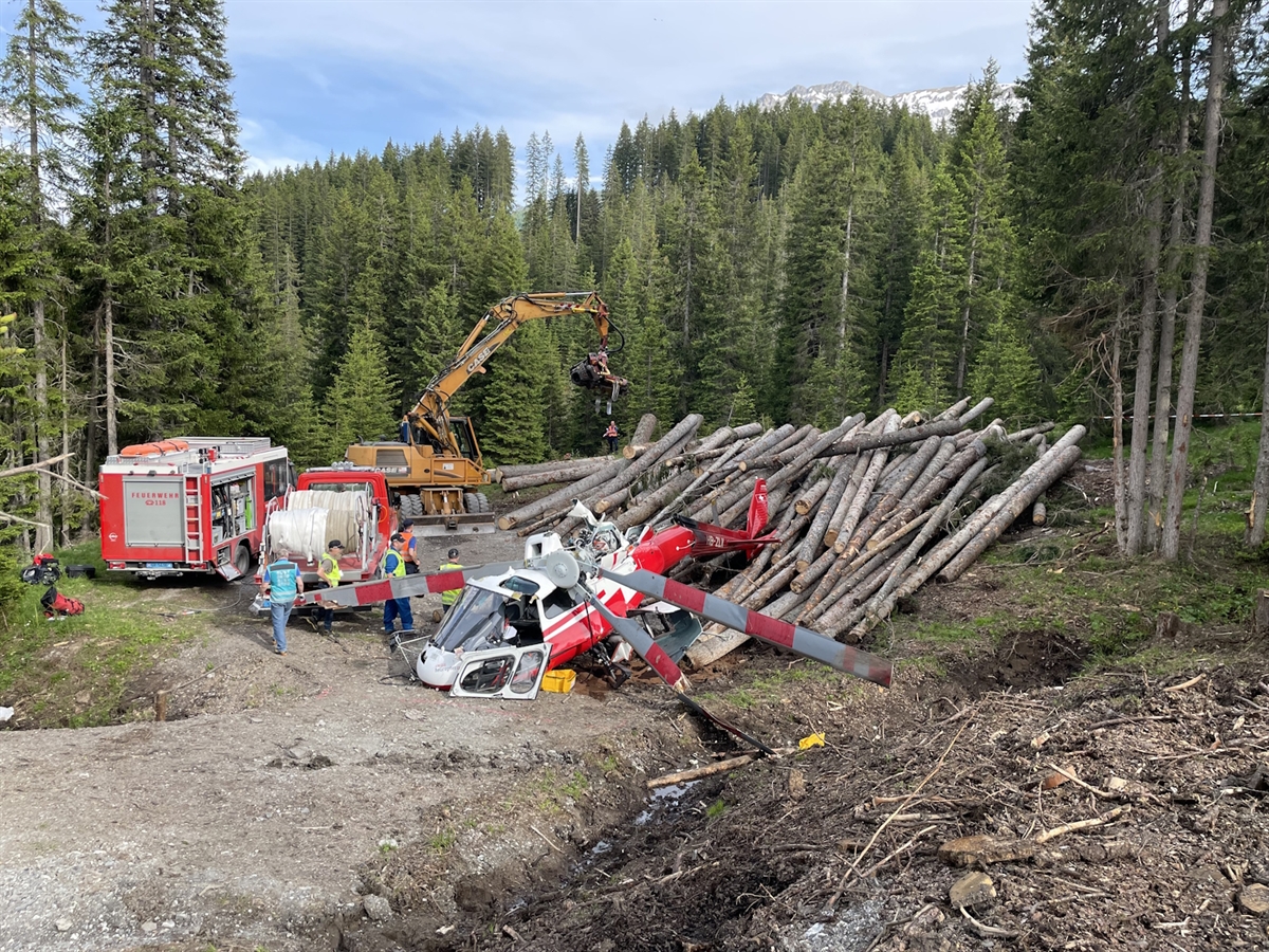 Der Helikopter liegt total beschädigt auf einem Holzlagerplatz. Daneben Einsatzkräfte. Im Hintergrund Wald und Horizont.
