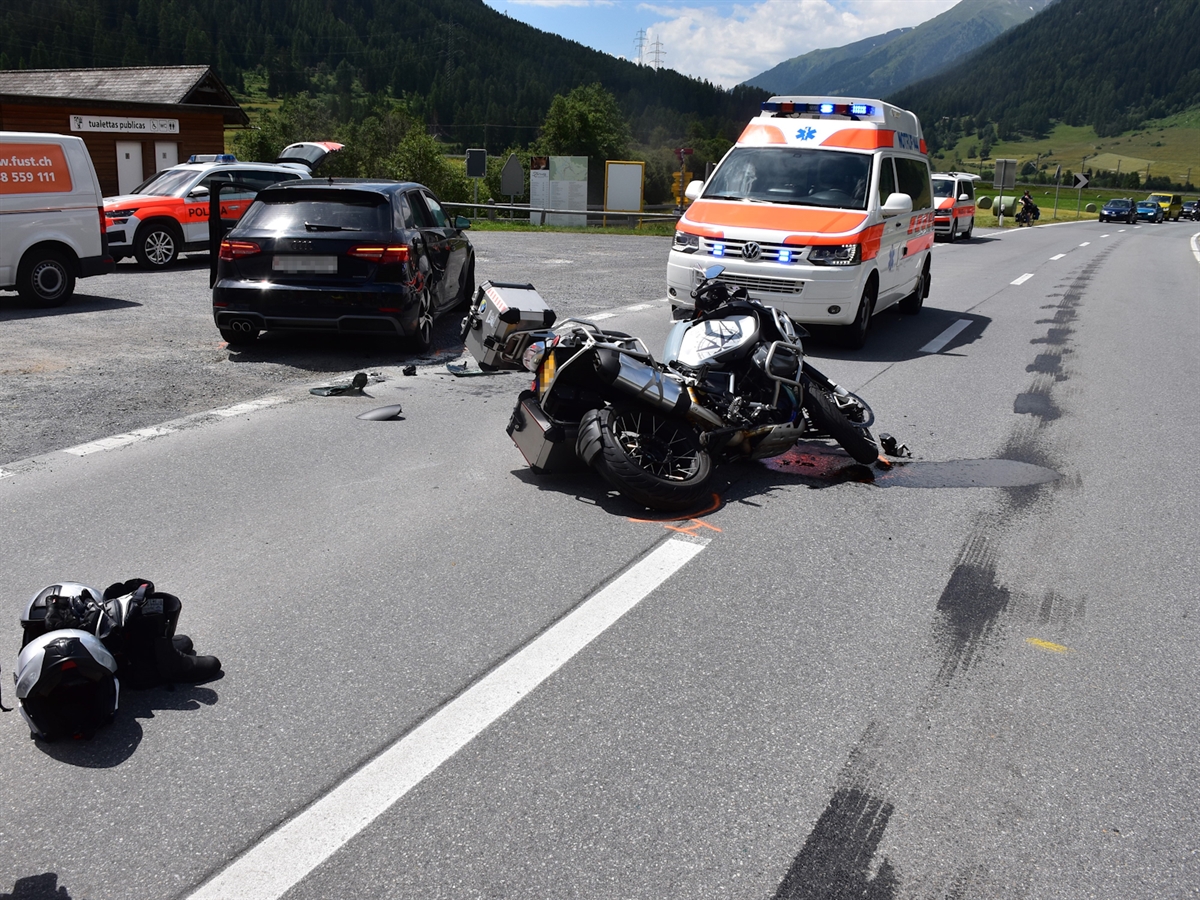 Das Motorrad liegt seitlich auf der Ofenbergstrasse. Links davon auf dem Parkplatz das Unfallauto. Hinter dem Töff steht ein Ambulanzfahrzeug sowie zwei Patrouillenfahrzeuge der Kantonspolizei Graubünden.