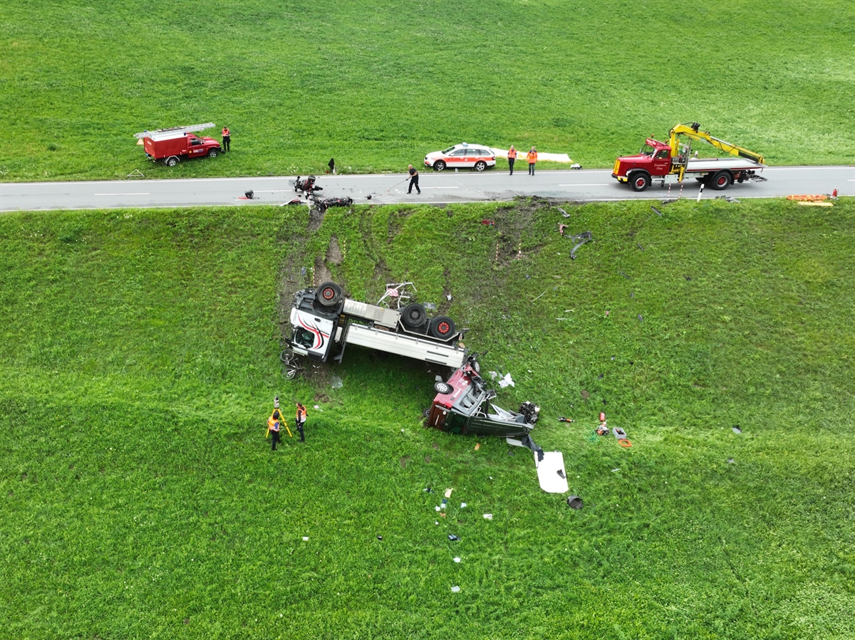 Die Unfallstelle mit dem gekippten Lastwagen und dem total beschädigten Wohnmotorwagen in der Böschung. Oberhalb dieser liegen zwei Motorräder auf der Strasse. Ein Kranwagen steht für die Bergung der Fahrzeuge bereit. Zwei Polizisten vermessen mit dem 3D-Scanner die Unfallstelle.