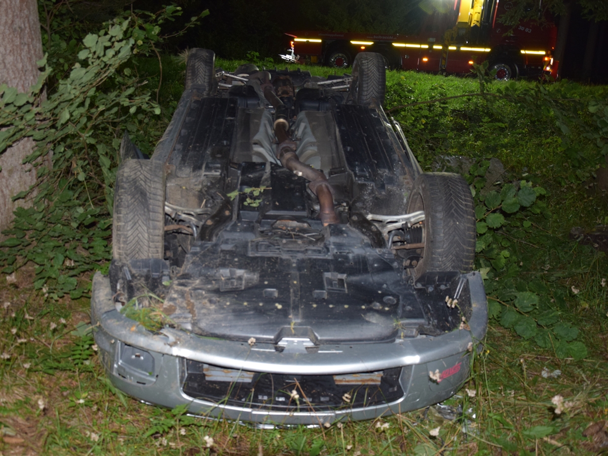 Das zwischen Bäumen auf dem Dach liegende Auto. Im Hintergrund die reflektierenden Streifen des Bergungsfahrzeuges des Abschleppdienstes.