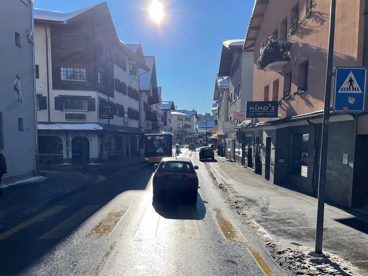 Die Hauptstrasse in Lenzerheide. Mitten im Bild das Auto mit welchem der Fussgänger angefahren wurde. Die Sonne scheint am oberen Bildrand und spiegelt sich auf der nassen Fahrbahn.