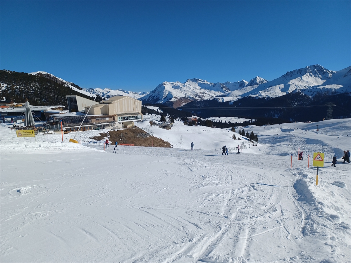 Die Piste führt auf einen Fussgängerweg zu. Links die Mittelstation. Im Hintergrund Bergpanorama mit blauem Himmel.