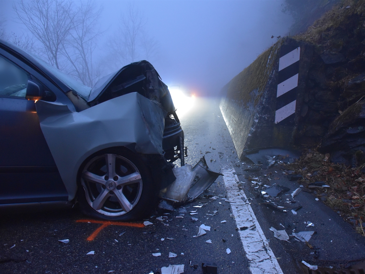 Die total beschädigte Front des Unfallautos neben dem Beginn der Stützmauer. Auf der Fahrbahn liegen viele Fahrzeugtrümmerteile.
