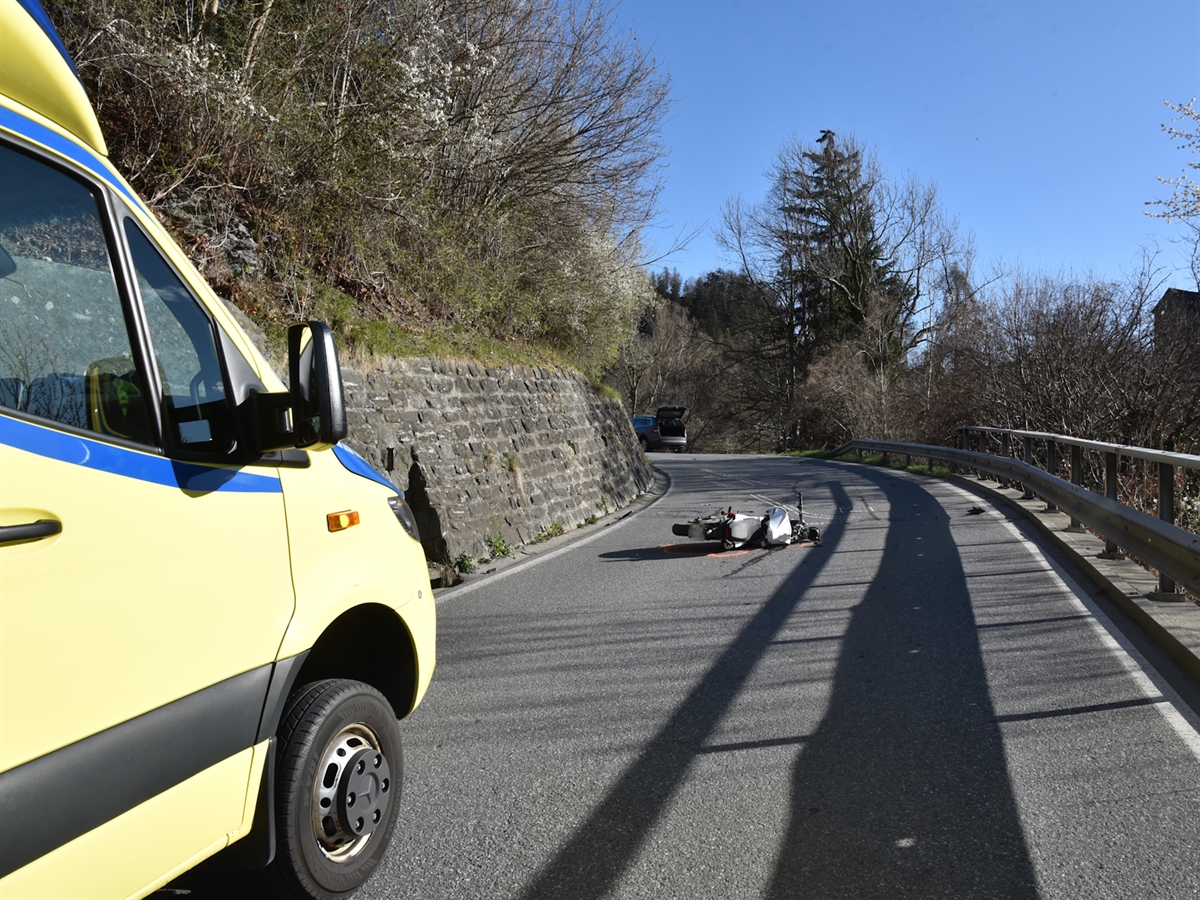 Am linken Bildrand die Front des Rettungswagens, in der Bildmitte liegt das Motorrad mitten auf der Fahrbahn. Im Hintergrund auf einem Ausstellplatz steht das Auto, in welches das Motorrad gerutscht ist. Das frühlingshafte Bild wird mit blauem Himmel abgerundet.