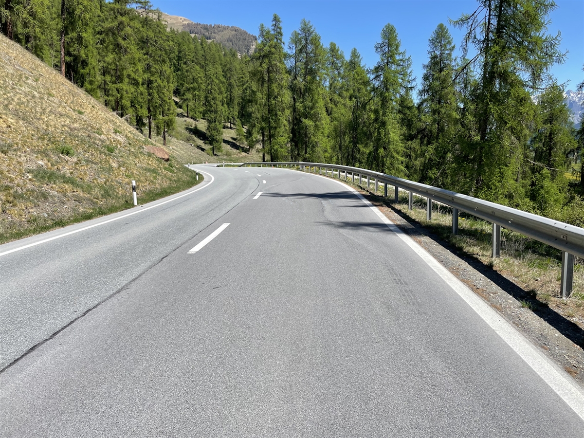 Der Verlauf der Engadinerstrasse mit der Linkskurve und rechtsseitigen Leitplanke. Der Engadinerwald wird mit strahlend blauem Himmel ergänzt.