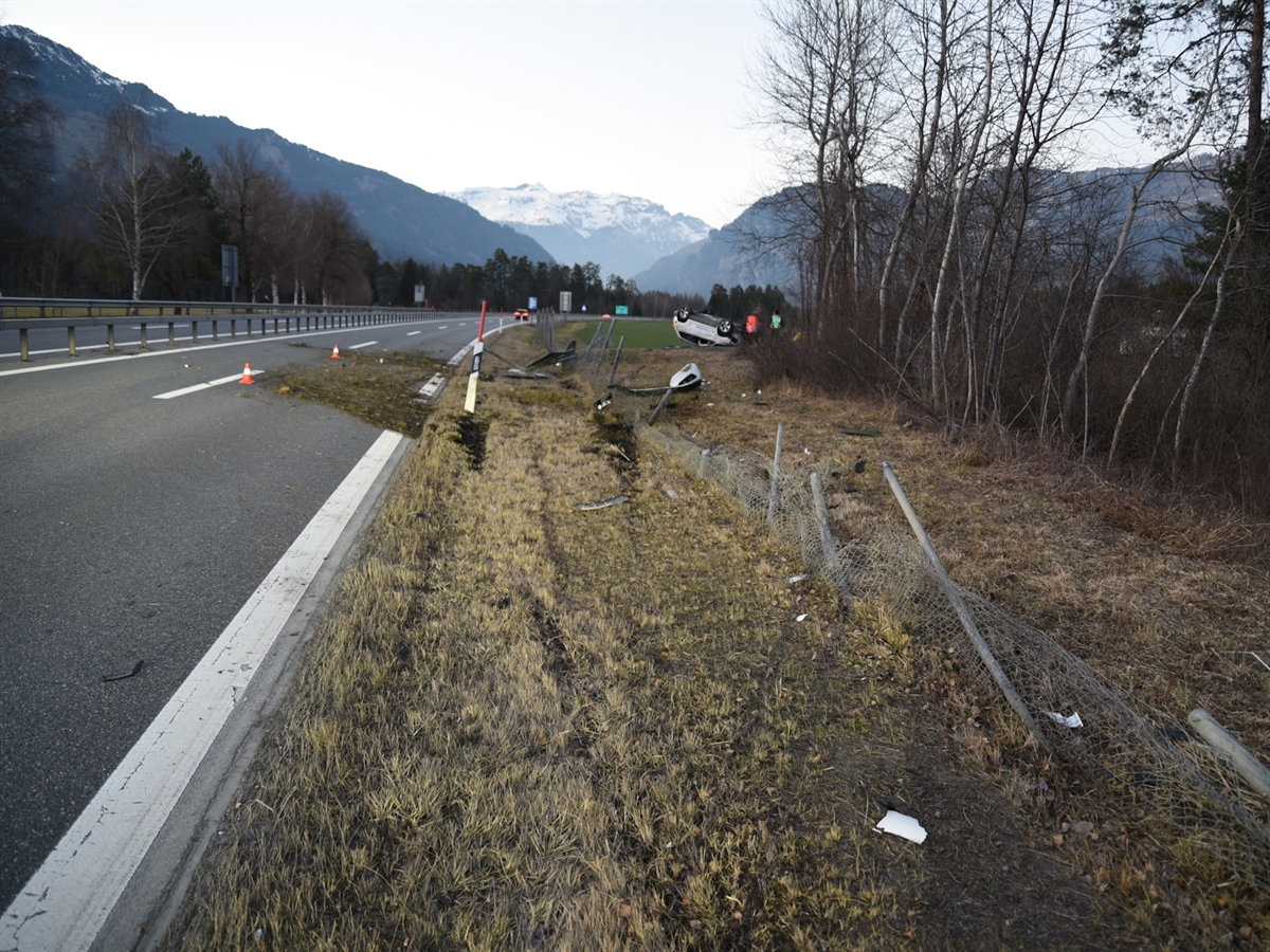 Im Vordergrund die Autostrasse und die rechtsseitige Wiesenböschung. Beschädigter Wildschutzzaun und Kollisionsspuren. Im Hintergrund das auf dem Dach liegende Auto.