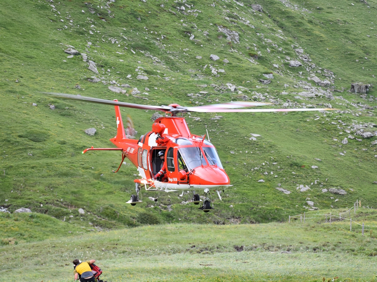 Der Helikopter der Rega beim Landeanflug. Zwei Personen knien am Boden.