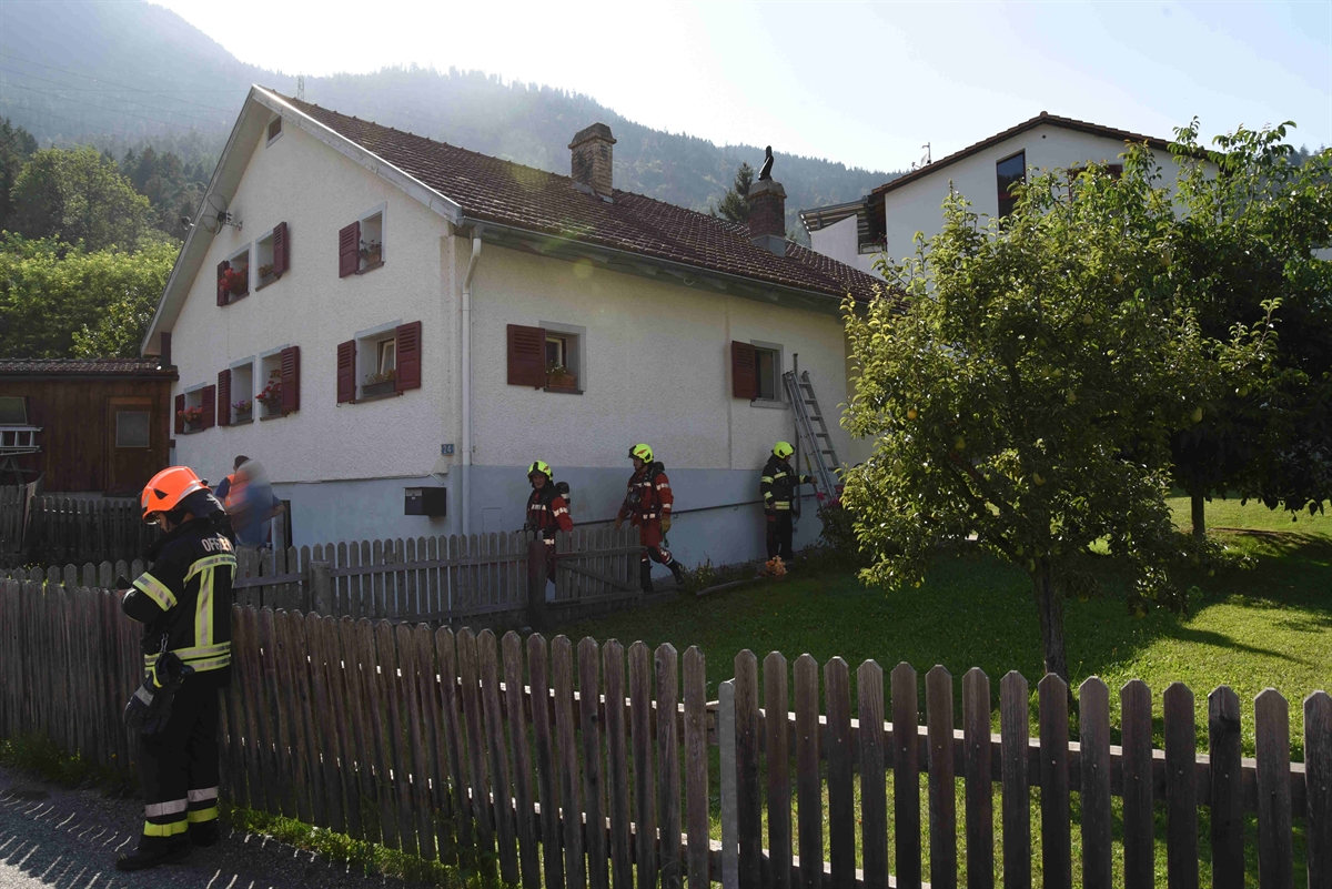 Älteres EFH inmitten von Grünfläche umzäunt mit einem Holzzaun und mehrere Feuerwehrleute stehen beim Haus und eine Leiter ist zu einem Fenster gestellt.