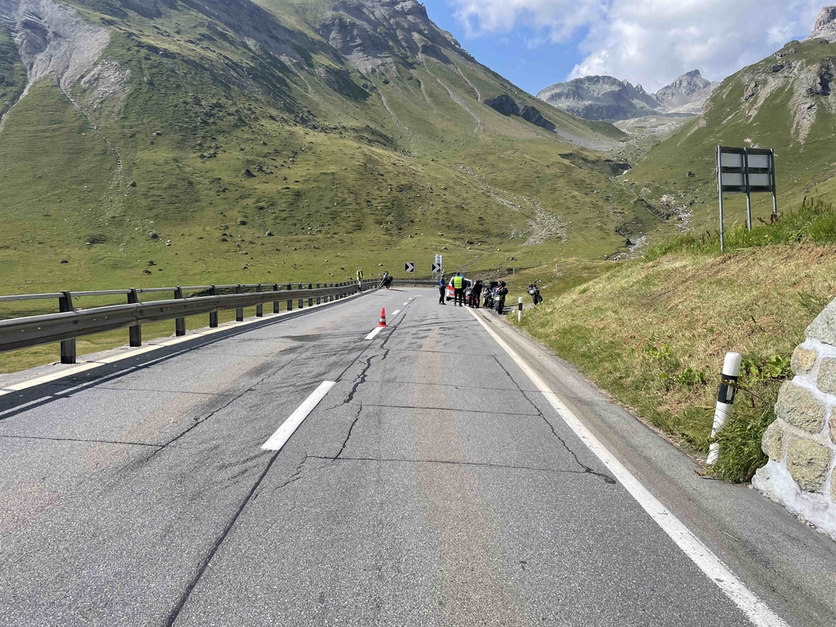 Beginn der Strasse im Kurvenausgang mit anschliessender Geraden. Im Hintergrund Rechts am Strassenrand die Motorradgruppe und der Polizist.