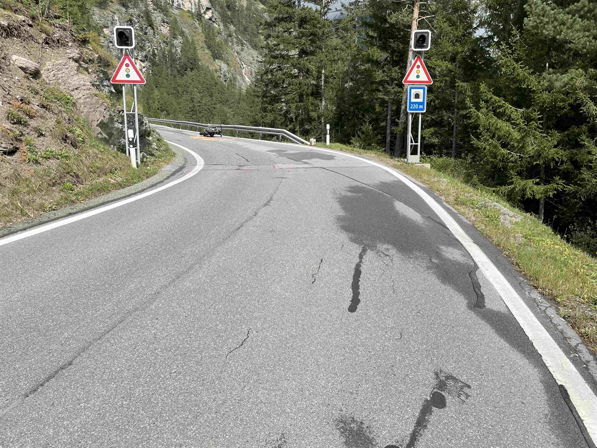 Strassenführung mit unübersichtlicher Linkskurve. Kurvenausgang liegt das Motorrad rechts am Boden. Links und rechts der Strasse Wald und Wieslandböschung. Kurveneingang ist beidseitig die Gefahrensignale Lichtsignalanlage und Hinweissignal Tunnel.
