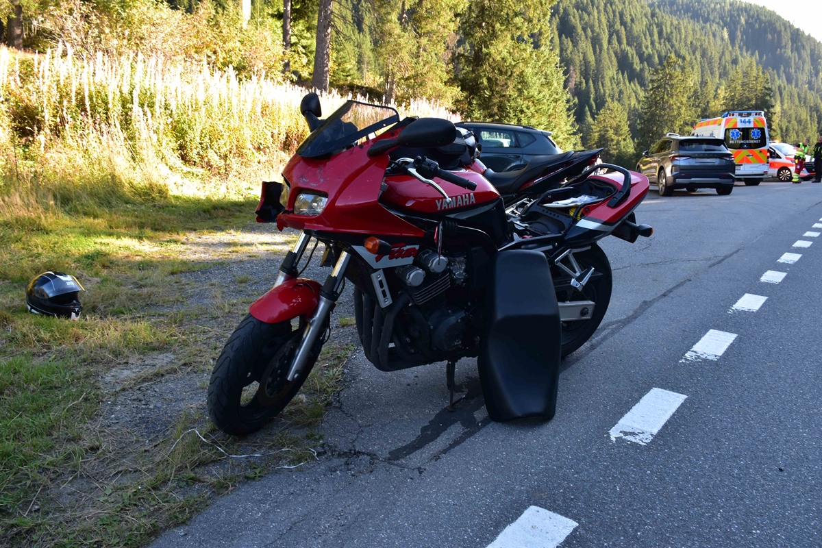 Das rote, beschädigte Motorrad steht am rechten Fahrbahnrand in Fahrtrichtung Klosters. Auf der rechtseitigen Wiese liegt ein Helm. Hinten im Bild stehen eine Ambulanz, Polizeifahrzeug und das im Unfall involvierte Auto.