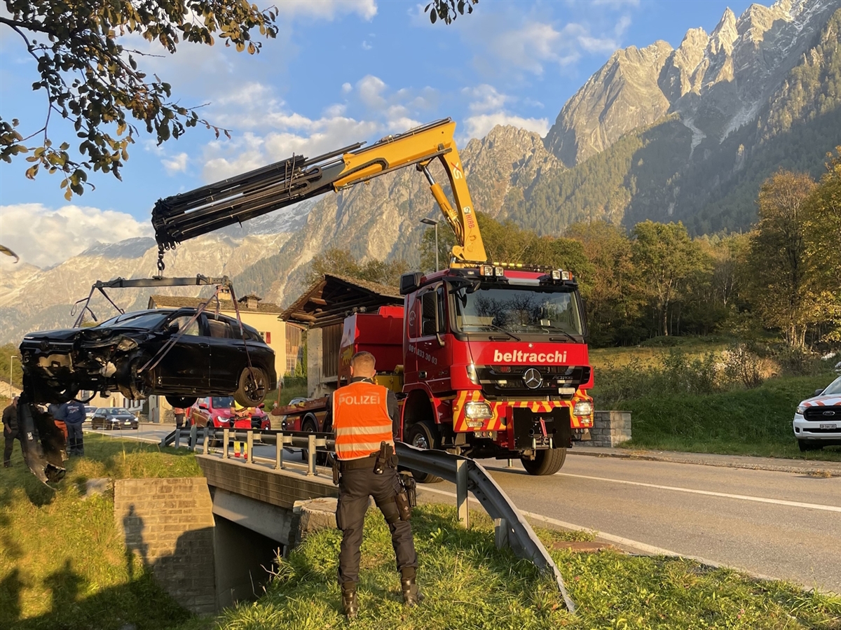 Ein Polizist der die Unfallstelle fotografiert sowie ein Bergungsfahrzeug, welches das Unfallauto anhebt. Im Hintergrund Berge die sich vom grösstenteils heiteren Horizont abgrenzen.