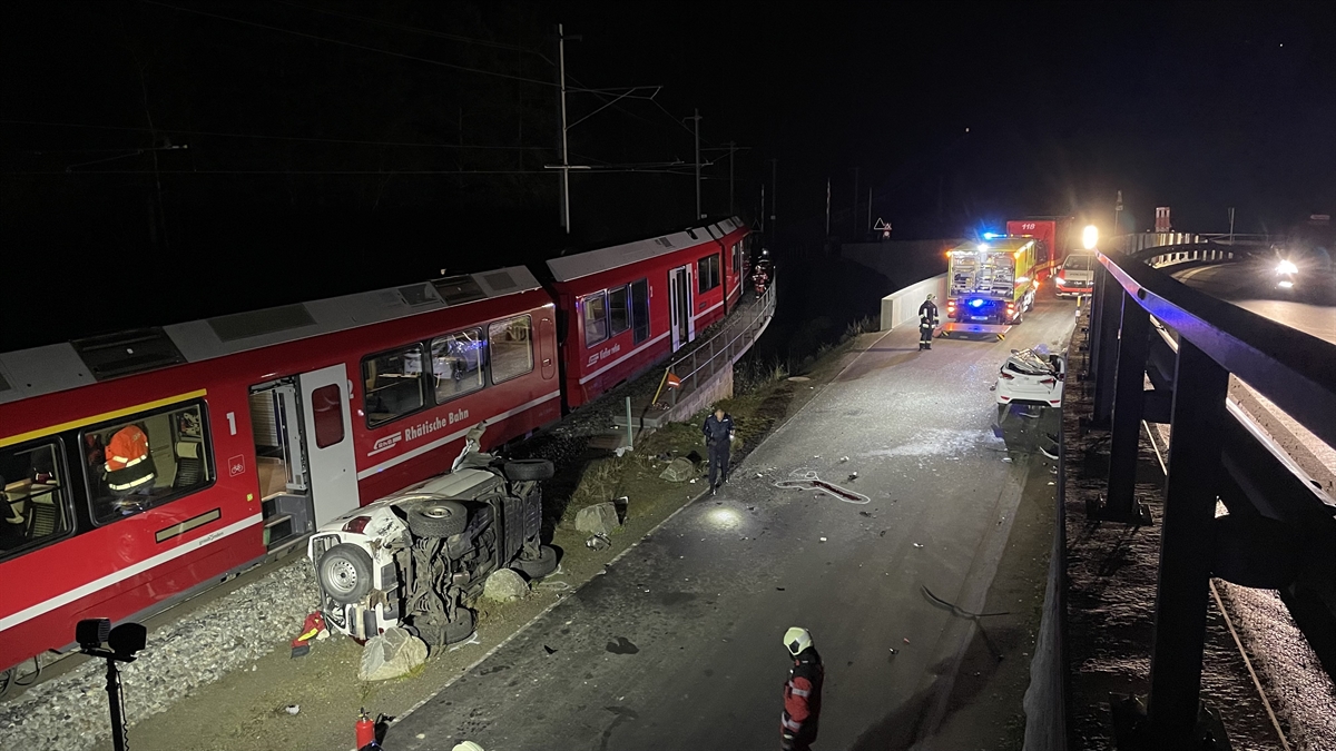 Unfallstelle in Miralago. Auf der linken Seite ein stehender Zug der RhB. Daneben ein auf der Seite liegender weisser Personenwagen. Im Hintergrund auf der rechten Seite unterhalb der Stützmauer ein weiteres totalbeschädigtes Auto mit eingedrücktem Dach. Im Hintergrund ist noch ein Fahrzeug der Feuerwehr zu erkennen.
