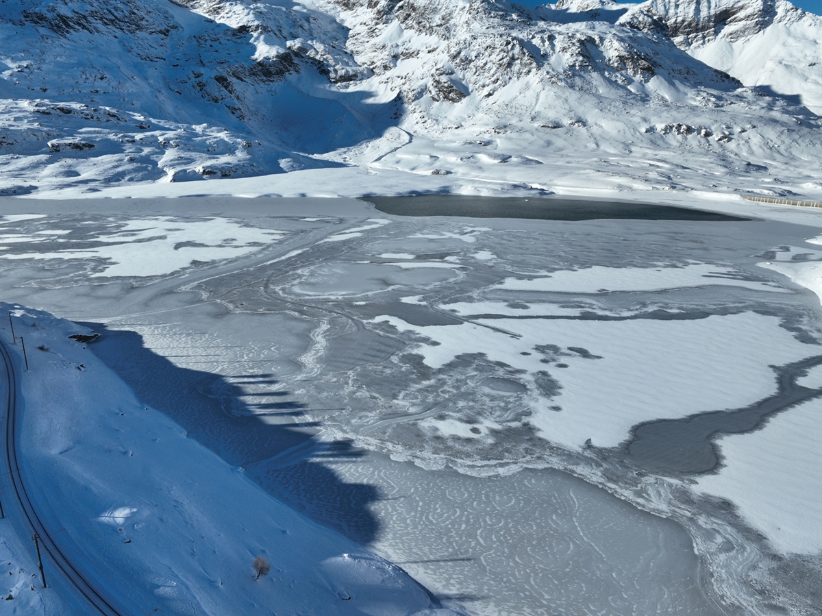 Der teilweise zugefrorene Lago Bianco am Tag danach. Durch die Sonneneinstrahlung werden die Schatten von Bergen und Hügeln auf den zugefrorenen See gezeichnet