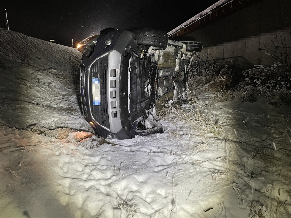 Das auf verschneitem Gelände auf der Beifahrerseite liegende Auto von vorne. Dahinter die zur Strasse ansteigende Böschung.