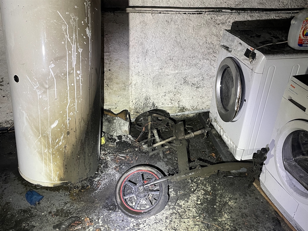 Ein verkohlter E-Scooter liegt zwischen einer Waschmaschine und einem Boiler. Russ und verbranntes Plastik zieren den gesamten Boden sowie die Wände der Waschküche