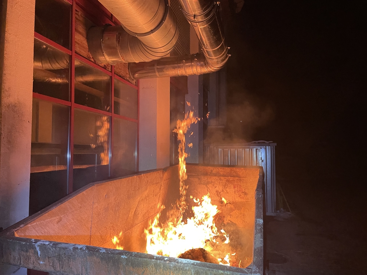 Flammen steigen aus einer Mulde empor. Über der Mulde Abluftrohre aus dem Gebäude. Das Feuer erhellt die Nacht mit warmem Licht.