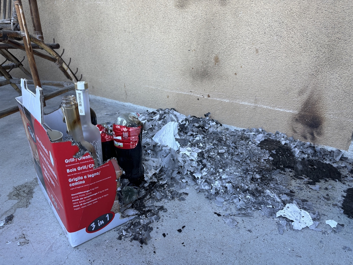Auf dem Betonboden liegt Asche und eine angesengte Kartonschachtel mit leeren PET-Flaschen, welche ebenfalls durch den Brand in Mitleidenschaft gezogen wurden.